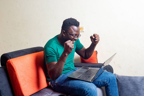 Nuori mies lasit istuu sohvalla kotona työskentelee kannettavan tietokoneen ja osoittaa voittavan ele. - Valokuva, kuva