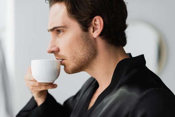 Side view of young man in robe holding coffee in bathroom  - 写真・画像