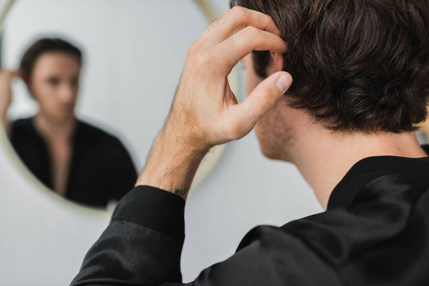 Young man in satin robe touching hair near blurred mirror at home  - Фото, зображення