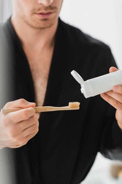 Cropped view of blurred man in black robe holding toothbrush and toothpaste in bathroom  - Foto, Imagen