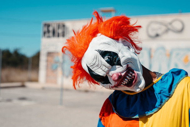Крупный план страшного злого клоуна, с рыжими волосами и в красочном костюме, строящего рожи, глядя на наблюдателя в одиноком месте на улице - Фото, изображение