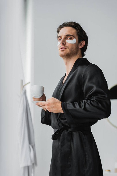Low angle view of man in silk robe with eye patches holding cup of coffee in bathroom  - Фото, зображення