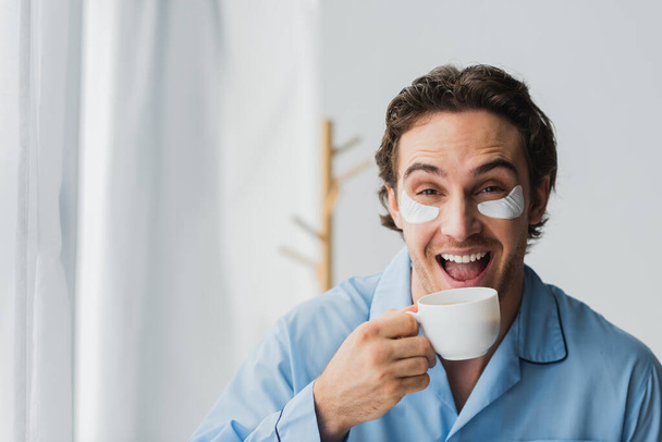Cheerful man in eye patches and pajama holding cup of coffee at home  - Fotografie, Obrázek
