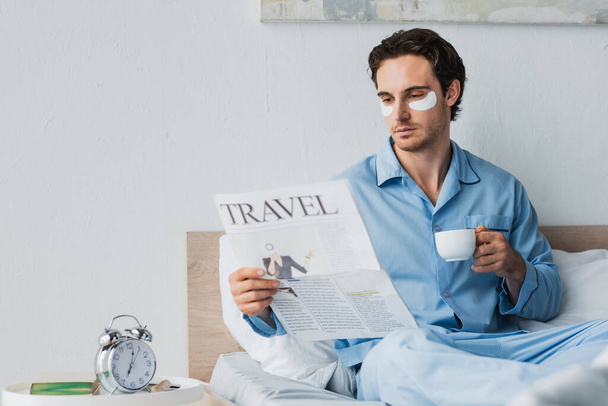 Άντρας με μπαλώματα στα μάτια κρατώντας καφέ και διαβάζοντας εφημερίδα στο κρεβάτι το πρωί  - Φωτογραφία, εικόνα