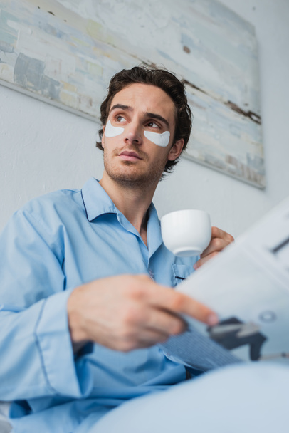 Tiefansicht des Mannes in Augenklappen und Pyjama mit Kaffeetasse und verschwommener Zeitung im Schlafzimmer  - Foto, Bild