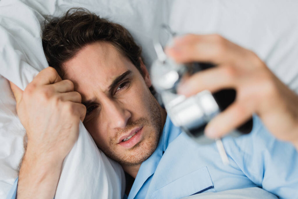 High angle view of upset man holding blurred alarm clock on bed  - Fotó, kép