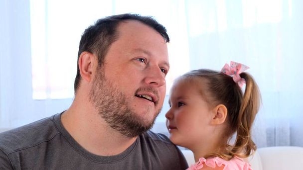 little daughter, 3 years old, tells her dad a secret, play together at home. Fathers day. Handsome young man at home with his little cute girl are having fun together. Fatherhood concept, happy - Фото, зображення