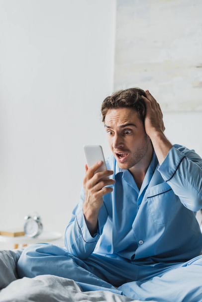 Scared man in pajama looking at smartphone on bed in morning  - Foto, Bild