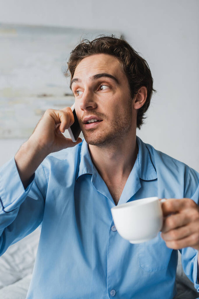 Muž s šálkem kávy mluví na smartphone v ložnici  - Fotografie, Obrázek