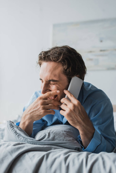 Cheerful man in pajama talking on smartphone on bed in morning  - Foto, Imagem