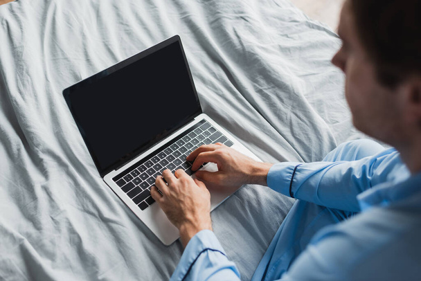 High angle view of blurred man in pajama using laptop with blank screen on bed  - Фото, зображення