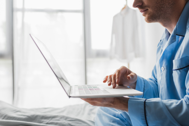 Vista recortada de un joven en pijama usando un portátil en la cama por la mañana  - Foto, Imagen