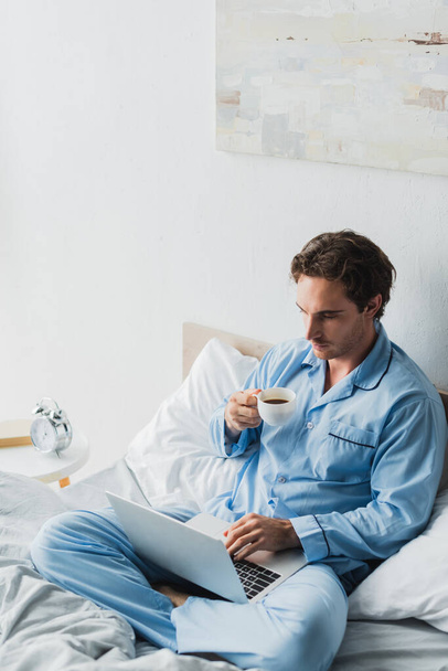 Фрилансер в пижаме с чашкой кофе во время использования ноутбука на кровати  - Фото, изображение