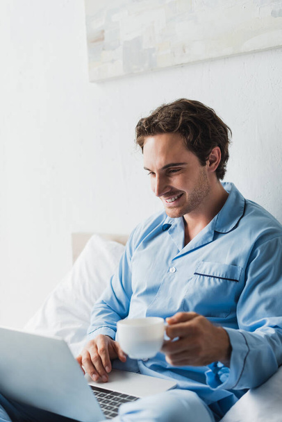 Smiling freelancer in pajama using laptop and holding blurred coffee cup on bed in morning  - Фото, зображення