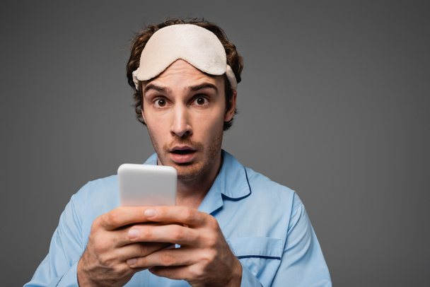 Shocked man in sleep mask and pajama holding smartphone and looking at camera isolated on grey  - Foto, Imagem