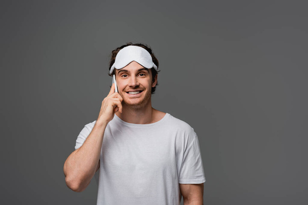 Smiling man in sleep mask and white t-shirt talking on smartphone isolated on grey  - Fotoğraf, Görsel