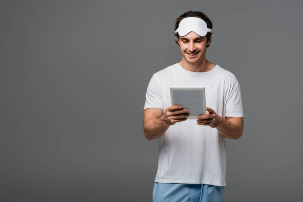 Homem sorridente em máscara de sono e pijama usando comprimido digital isolado em cinza  - Foto, Imagem