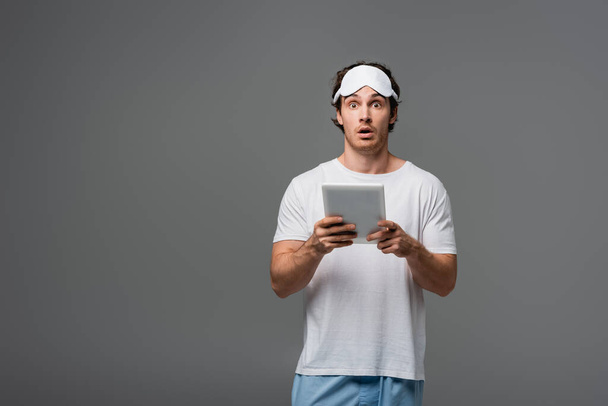 Hombre sorprendido en la máscara del sueño y pijama usando tableta digital aislada en gris  - Foto, Imagen