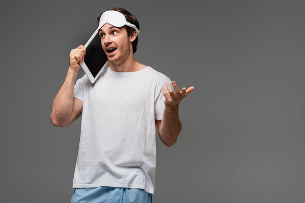 Junger Mann in Schlafmaske und Pyjama spricht auf digitalem Tablet isoliert auf grau  - Foto, Bild