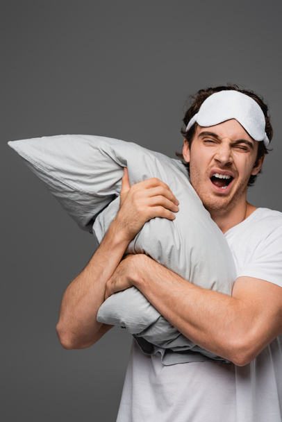 Человек в маске для сна зевает, держа в руках подушку, изолированную от серых  - Фото, изображение