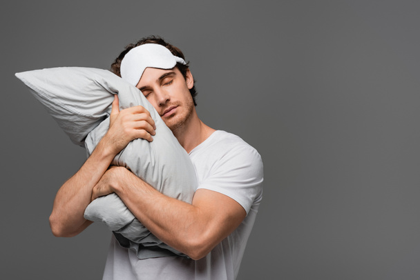 Young man in sleep mask hugging pillow isolated on grey  - Photo, Image