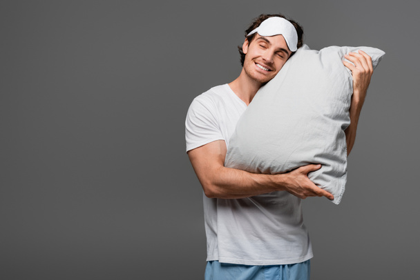 Young man in sleep mask hugging pillow isolated on grey  - Foto, imagen