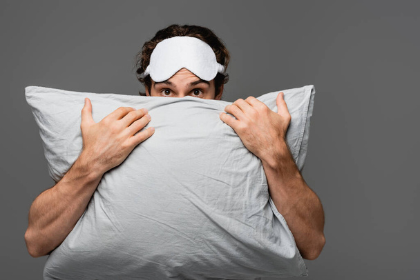 Jovem com máscara de dormir cobrindo rosto com travesseiro isolado em cinza  - Foto, Imagem