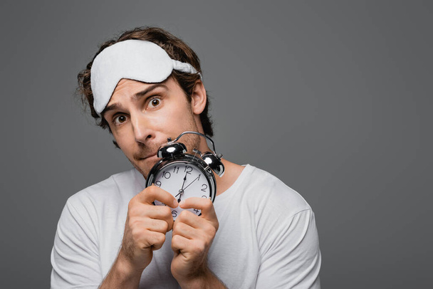 Tensed man in sleep mask holding alarm clock isolated on grey  - Фото, изображение