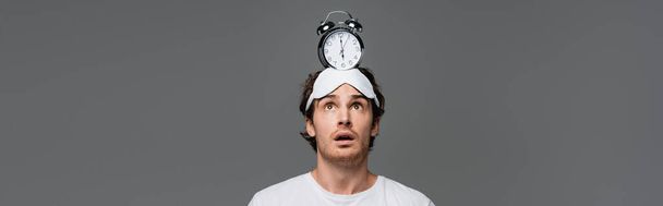 Verbaasde man met slaapmasker kijkend naar wekker op hoofd geïsoleerd op grijs, spandoek  - Foto, afbeelding