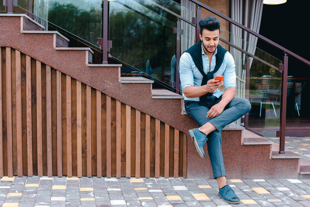 Atractivo joven árabe de pie con la maleta en la mano y posando para la cámara, poner un pie antes que el otro y se basa en las escaleras cerca del restaurante al aire libre. - Foto, imagen