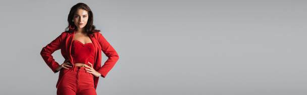 Kırmızı takım elbiseli, küpeli, elleri kalçasında poz veren, gri pankartta izole genç bir kadın. - Fotoğraf, Görsel