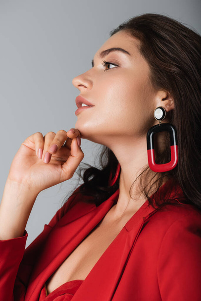 side view of trendy woman in red suit and earring posing isolated on grey - Fotografie, Obrázek