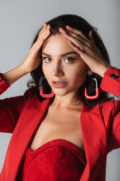 young trendy woman in red suit and earrings looking at camera isolated on grey - Фото, изображение