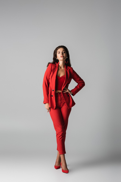 full length of young confident woman in red heels and suit posing with hand on hip on grey - Фото, изображение