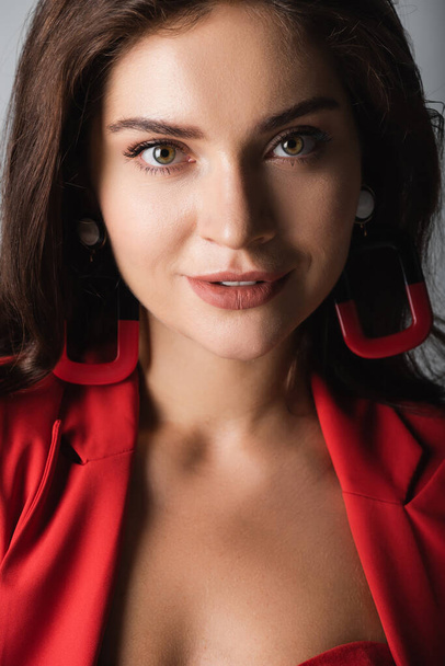 pleased young woman in red suit and earrings looking at camera isolated on grey - Fotoğraf, Görsel