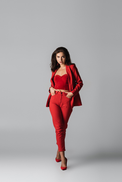 full length of young stylish woman in red heels and suit posing with hands in pockets on grey - Foto, Imagem