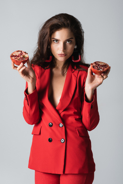 stylish woman in trendy suit holding red pomegranate halves isolated on grey - Foto, Imagem