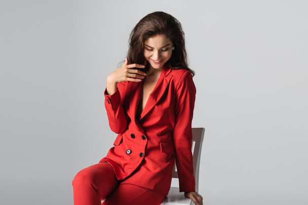cheerful young woman in red suit posing while sitting on white chair isolated on grey - Foto, afbeelding