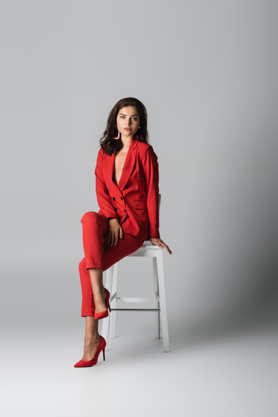 full length of trendy woman in red stylish suit posing while sitting on white chair on grey - Fotoğraf, Görsel