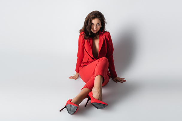 comprimento total da mulher bonita em terno vermelho posando enquanto sentado no cinza - Foto, Imagem