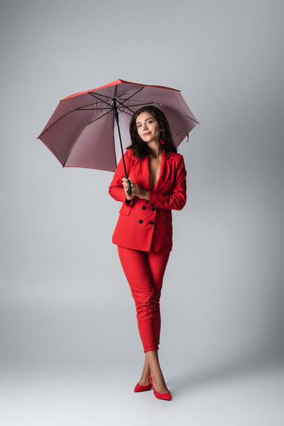volledige lengte van brunette vrouw in rood pak staan onder paraplu op grijs - Foto, afbeelding