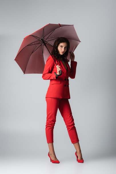 full length of young woman in red suit standing under umbrella on grey - Fotografie, Obrázek