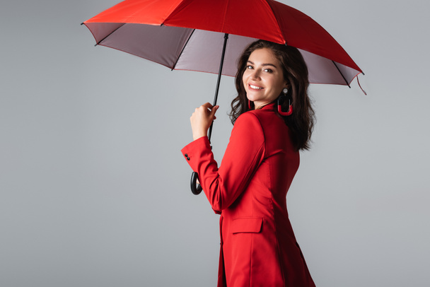 happy young woman in red suit standing under umbrella isolated on grey - Φωτογραφία, εικόνα