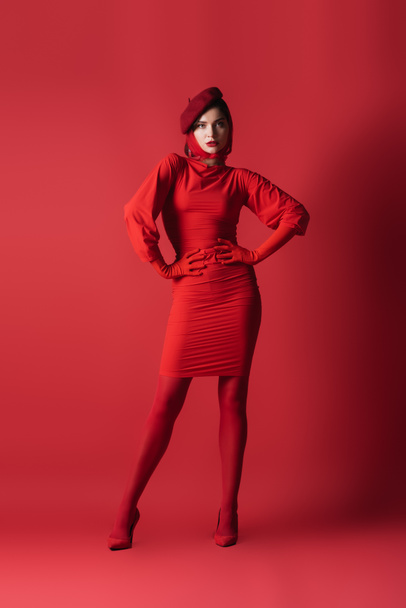 full length of young brunette woman in beret posing with hands on hips on red - Fotografie, Obrázek