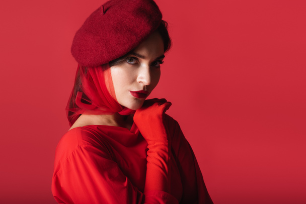 elegante mujer joven en boina y guante mirando a la cámara aislada en rojo - Foto, imagen