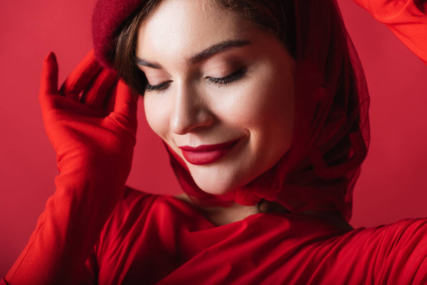 close up of smiling young woman adjusting beret isolated on red - Photo, Image