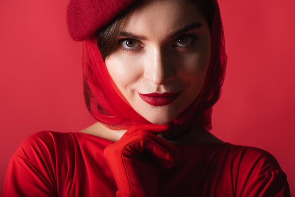 cheerful young woman in glove, headscarf and beret isolated on red - Photo, Image