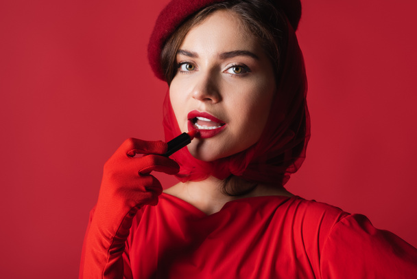 young woman in glove, headscarf and beret applying lipstick isolated on red - Foto, afbeelding