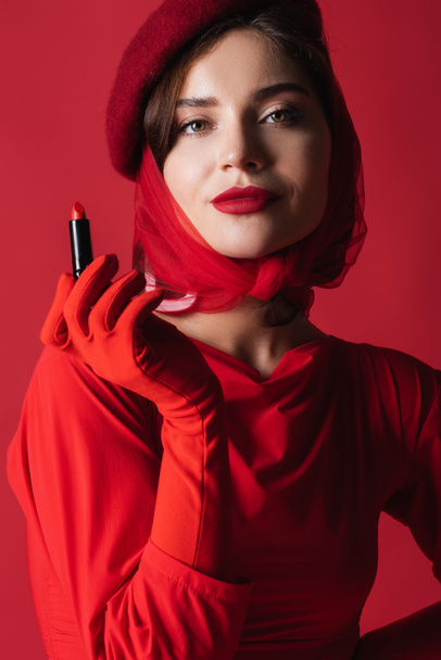 young woman in headscarf and beret holding lipstick isolated on red - 写真・画像