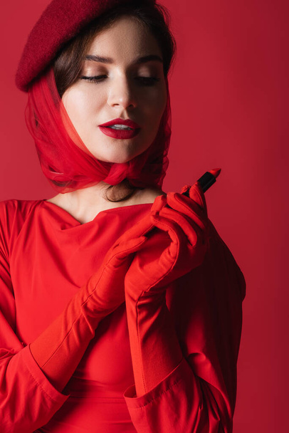 young woman in headscarf and beret holding lipstick isolated on red - Foto, immagini
